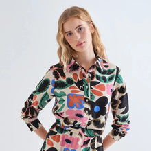 Load image into Gallery viewer, A woman modeling a long-sleeve collared shirt dress with a colorful abstract floral print on a light beige background, set against a plain light gray studio backdrop.
