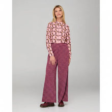 Load image into Gallery viewer, A woman modeling a pink long-sleeve blouse with a black and orange abstract pattern, paired with magenta and white wide-leg trousers, against a light gray studio background.
