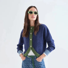 Load image into Gallery viewer, A woman modeling a navy blue knit cardigan with a green button placket, paired with blue patterned pants, a white top, and green sunglasses, against a plain white studio background.
